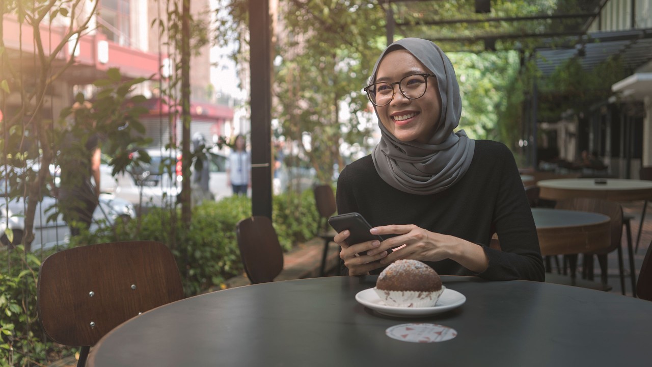 A young woman is happily sitting in a cafe, surrounding with lots of plants; imaged used for HSBC Malaysia Amanah financing concept 101 page. 